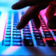 Fingers typing on laptop keyboard at night, illuminated by red, blue, and pink lights