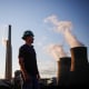 A worker stands in the coal yard, smoke stacks in the distance behind him blow smoke plumes into the sky