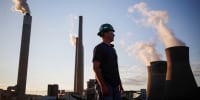A worker stands in the coal yard, smoke stacks in the distance behind him blow smoke plumes into the sky